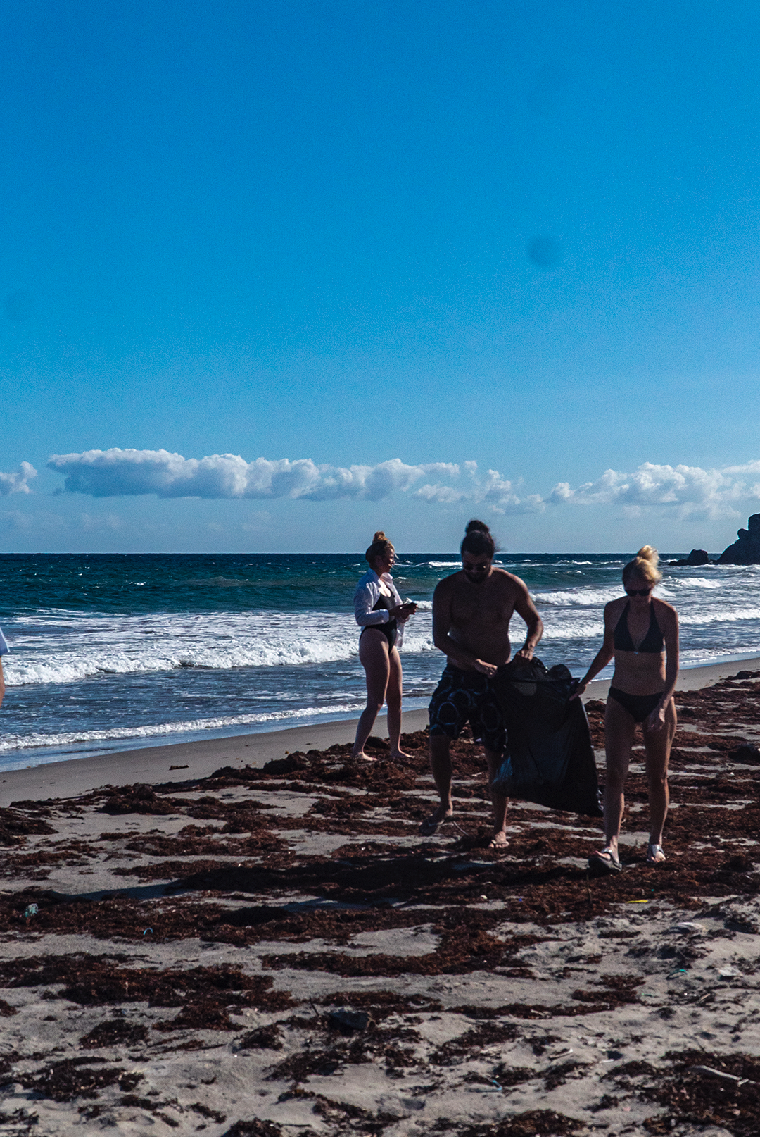 Seb Jemmett, Ghislaine Jemmett, and Susanna Tweedie-Hughes, the founding members of NAUTRA, participate in a beach cleanup, collecting debris along the shoreline under a bright blue sky. The ocean waves crash in the background as they work together to care for the environment.