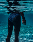 A woman stands partially submerged, with only her lower body visible, wearing the NAUTRA bikini bottoms in the Eagle Ray print. The ocean water shimmers around her.