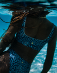 A woman wearing the Eagle Ray Print Scoop Neck Sports Bra Bikini Top from NAUTRA floats gracefully underwater, her hair flowing behind her as sunlight dances on the surface.