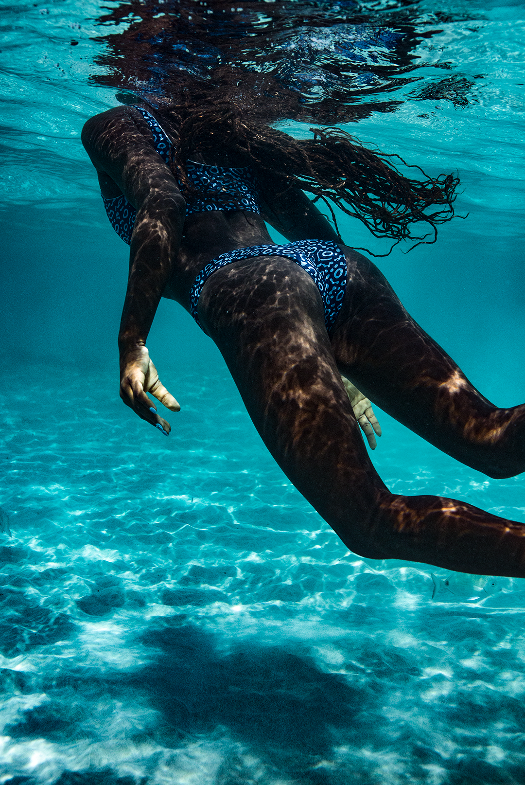 A woman gracefully swims underwater, wearing the NAUTRA bikini bottoms in the Eagle Ray print. Her long hair flows behind her as sunlight filters through the water, creating a mesmerizing pattern.