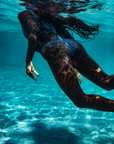 A woman gracefully swims underwater, wearing the NAUTRA bikini bottoms in the Eagle Ray print. Her long hair flows behind her as sunlight filters through the water, creating a mesmerizing pattern.