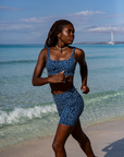 he woman runs along the beach in the Eagle Ray Print Scoop Neck Sports Bra Bikini Top from NAUTRA, her confident stride framed by the turquoise ocean and clear skies.