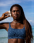 The woman holds a conch shell to her ear, listening intently to the sound of the ocean, wearing the Eagle Ray Print Sports Bra Bikini Top from NAUTRA.