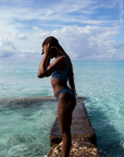 A woman adjusts her snorkel mask while standing on a rocky platform over clear ocean water, wearing the NAUTRA Eagle Ray Print High Hike Bikini Bottoms.