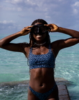 A woman smiles while adjusting her snorkel mask, standing near the ocean's edge in the NAUTRA Eagle Ray Print High Hike Bikini Bottoms. The water glistens in the sunlight.