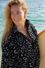 A woman is walking barefoot along a sandy beach, holding a surfboard in one hand and smiling towards the camera. She is wearing the NAUTRA EAGLE RAY Print fleece, black wetsuit leggings with turquoise accents, and has wet hair from the ocean.