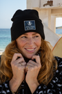 A model with long red hair adjusts a black NAUTRA Recycled Beanie featuring the brand's logo and slogan, 'Be the Wave.' She is also wearing the Eagle Ray print fleece, with its striking black and white pattern. The background shows a sunny beach scene with a surfboard and wooden structure.