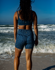 A woman stands facing the ocean in the NAUTRA Eagle Ray Print Biker Shorts. Her back is to the camera, and the waves crash on the shore under a bright blue sky.