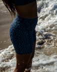 Close-up of a woman’s back, wearing the NAUTRA Eagle Ray Print Biker Shorts and matching top. The seafoam swirls around her legs as she faces the waves.