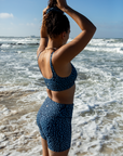 A woman ties her hair into a bun on the beach while wearing the NAUTRA Eagle Ray Print Biker Shorts. The ocean waves crash in the background, and her focus is on the task.