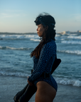 A woman holds diving fins while standing on the beach in the NAUTRA Eagle Ray Print Long-Sleeve One-Piece Swimsuit. The waves crash behind her as she gazes out to sea, soaking in the ocean breeze. 