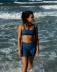 A woman walks along the shore, wearing the NAUTRA Eagle Ray Print Biker Shorts. The waves crash around her legs as she enjoys the beach.