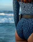 A close-up of a woman’s back wearing the NAUTRA Eagle Ray Print Long-Sleeve One-Piece Swimsuit with a snorkel belt. Her red hair contrasts with the patterned swimsuit as ocean waves roll in the background.