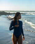 A woman walks along the shoreline in the NAUTRA Eagle Ray Print Long-Sleeve One-Piece Swimsuit, with her wet hair flowing behind her. She enjoys the ocean breeze, with waves gently rolling in.