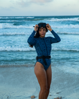 A woman smiles while standing on a beach, wearing the NAUTRA Eagle Ray Print Long-Sleeve One-Piece Swimsuit. The waves crash behind them, and they hold a snorkel belt, enjoying the sunset.