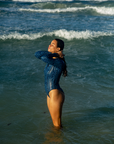 A woman stands knee-deep in the ocean, wearing the NAUTRA Eagle Ray Print Long-Sleeve One-Piece Swimsuit. She tilts her head back, enjoying the sunlight, while waves surround her.