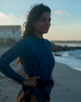 A woman smiles while standing on a beach, wearing the NAUTRA Eagle Ray Print Long-Sleeve One-Piece Swimsuit. The waves crash behind them, and they hold a snorkel belt, enjoying the sunset.