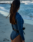 A woman faces the ocean, wearing the NAUTRA Eagle Ray Print Long-Sleeve One-Piece Swimsuit with a snorkel belt. Her wet hair flows in the wind as she looks towards the waves.