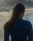 A close-up of a woman’s back wearing the NAUTRA Eagle Ray Print Long-Sleeve One-Piece Swimsuit with a snorkel belt, standing near the shoreline. The patterned suit contrasts with the foamy waves.