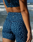 Close-up of a woman wearing the NAUTRA Eagle Ray Print Biker Shorts, facing the ocean. The detailed pattern is highlighted under the sunlight.