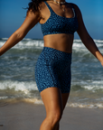 A woman stands with her arms outstretched on a sandy beach, wearing the NAUTRA Eagle Ray Print Biker Shorts. The ocean waves roll in behind her.