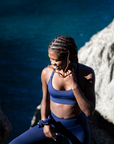 A Black female athlete in a Bluefin Navy yoga outfit from NAUTRA adjusts her hair while standing on a rugged coast, her thoughtful expression complemented by the expansive blue ocean in the background.