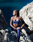 A Black woman in a Bluefin Navy athletic set from NAUTRA poses thoughtfully on a rocky coastline, looking into the distance with her hand resting on her bent knee, embodying a moment of contemplation.