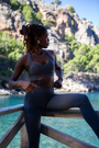 A Black female athlete sits on a wooden railing in the MINKE Grey Set from NAUTRA, overlooking a stunning sea scene. She appears thoughtful and composed, with the lush greenery and rocky landscape framing her.