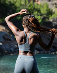 Rear view of a Black woman standing near a serene river, wearing the MINKE Grey Set from NAUTRA. She holds her braided hair up, showcasing the sports bra's back design against a scenic backdrop of forest and water.