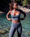 Fitness-focused Black woman in a MINKE Grey Set from NAUTRA stands by the water, preparing for a workout. Her intense gaze and strong posture are set against the natural beauty of a calm, clear bay.