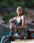 A woman sitting on a seaside dock, laughing joyfully while holding her knees close to her chest, wearing grey and black striped Wahoo leggings and a sports bra from NAUTRA.