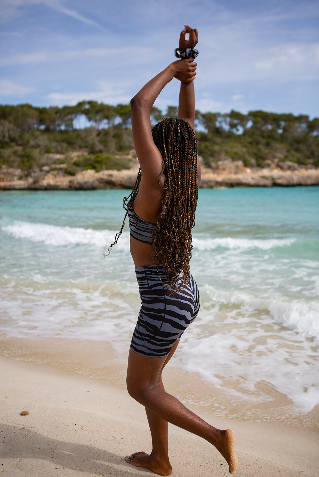 A woman in NAUTRA's grey and black striped Wahoo biker shorts and sports bra stands on a sandy beach with her arms stretched overhead, facing the ocean, with a green, forested area in the background.