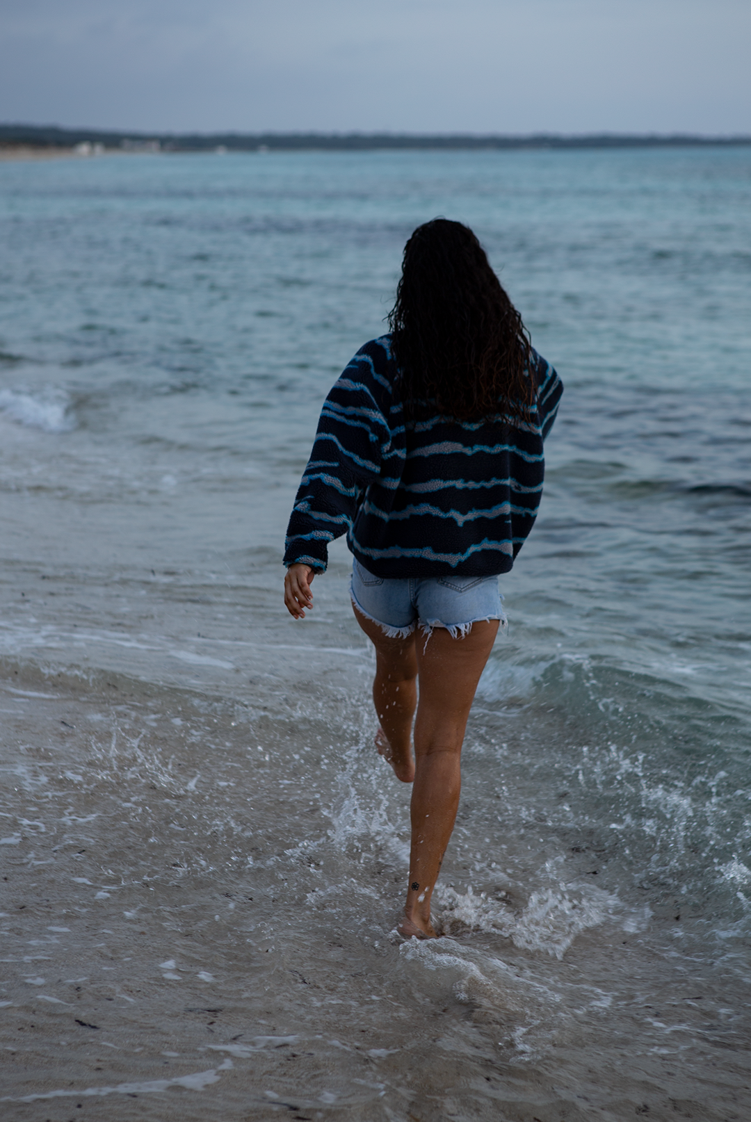 A woman on a sandy beach wearing the NAUTRA WAHOO Fleece, featuring vibrant blue tones and jagged stripes. Her hair is wet from the ocean, and she exudes a relaxed and cozy vibe, embracing the warmth of the fleece after a swim. The sea and sky create a serene backdrop.