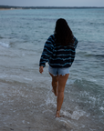 A woman on a sandy beach wearing the NAUTRA WAHOO Fleece, featuring vibrant blue tones and jagged stripes. Her hair is wet from the ocean, and she exudes a relaxed and cozy vibe, embracing the warmth of the fleece after a swim. The sea and sky create a serene backdrop.