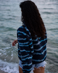 A woman on a sandy beach wearing the NAUTRA WAHOO Fleece, featuring vibrant blue tones and jagged stripes. Her hair is wet from the ocean, and she exudes a relaxed and cozy vibe, embracing the warmth of the fleece after a swim. The sea and sky create a serene backdrop.