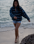 A woman on a sandy beach wearing the NAUTRA WAHOO Fleece, featuring vibrant blue tones and jagged stripes. Her hair is wet from the ocean, and she exudes a relaxed and cozy vibe, embracing the warmth of the fleece after a swim. The sea and sky create a serene backdrop.