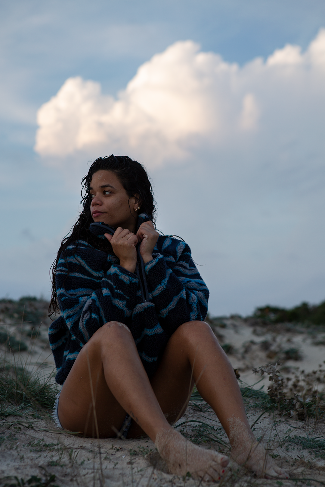 A woman on a sandy beach wearing the NAUTRA WAHOO Fleece, featuring vibrant blue tones and jagged stripes. Her hair is wet from the ocean, and she exudes a relaxed and cozy vibe, embracing the warmth of the fleece after a swim. The sea and sky create a serene backdrop.