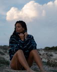 A woman on a sandy beach wearing the NAUTRA WAHOO Fleece, featuring vibrant blue tones and jagged stripes. Her hair is wet from the ocean, and she exudes a relaxed and cozy vibe, embracing the warmth of the fleece after a swim. The sea and sky create a serene backdrop.