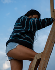 A woman on a sandy beach wearing the NAUTRA WAHOO Fleece, featuring vibrant blue tones and jagged stripes. Her hair is wet from the ocean, and she exudes a relaxed and cozy vibe, embracing the warmth of the fleece after a swim. The sea and sky create a serene backdrop.