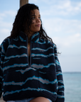 A woman on a sandy beach wearing the NAUTRA WAHOO Fleece, featuring vibrant blue tones and jagged stripes. Her hair is wet from the ocean, and she exudes a relaxed and cozy vibe, embracing the warmth of the fleece after a swim. The sea and sky create a serene backdrop.
