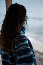 A woman on a sandy beach wearing the NAUTRA WAHOO Fleece, featuring vibrant blue tones and jagged stripes. Her hair is wet from the ocean, and she exudes a relaxed and cozy vibe, embracing the warmth of the fleece after a swim. The sea and sky create a serene backdrop.