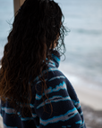 A woman on a sandy beach wearing the NAUTRA WAHOO Fleece, featuring vibrant blue tones and jagged stripes. Her hair is wet from the ocean, and she exudes a relaxed and cozy vibe, embracing the warmth of the fleece after a swim. The sea and sky create a serene backdrop.