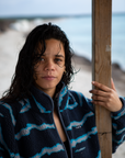 A woman on a sandy beach wearing the NAUTRA WAHOO Fleece, featuring vibrant blue tones and jagged stripes. Her hair is wet from the ocean, and she exudes a relaxed and cozy vibe, embracing the warmth of the fleece after a swim. The sea and sky create a serene backdrop.