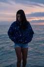 A woman stands on a sandy beach wearing the NAUTRA WHALE SHARK Fleece, designed with a pattern inspired by whale sharks. Her wet hair glistens as she gazes at a vibrant pink sunset, creating a tranquil and reflective atmosphere. The fleece keeps her warm after a swim, with the ocean and sky blending into a serene backdrop.